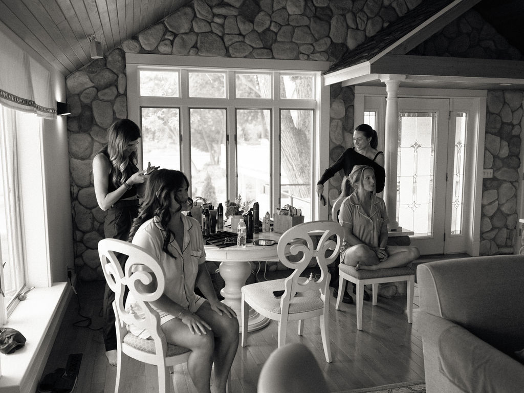 Black and white candid photo of a bride and her bridesmaid getting hair and makeup done.